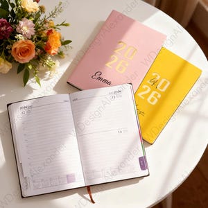 May include: An open 2026 diary with lined pages and a brown ribbon, alongside two closed planners in pink and yellow. The pink planner has the name "Emma" and the year "2026" in gold. The yellow planner also has the year "2026" in gold.