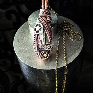 Puede incluir: Un colgante de estilo steampunk con envoltura de alambre de cobre y pequeños detalles de engranajes. El colgante presenta un centro claro y rectangular y está suspendido de una delicada cadena dorada. El diseño tiene una estética vintage e industrial.