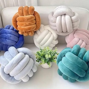 May include: A collection of knot-shaped decorative pillows in various colors, including orange, blue, white, gray, pink, and teal. The pillows are made of a soft, plush material and are arranged on a white surface.