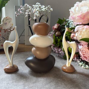 May include: A decorative vase composed of stacked, pebble-shaped elements in brown and white tones, filled with white flowers. Two deer figurines in white and brown are positioned nearby. The setting is a neutral surface with floral arrangements.