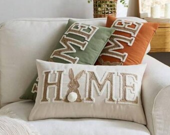 May include: A collection of decorative throw pillows on a white sofa. The pillows feature the word "HOME" with a bunny design and the word "ME" in a textured, embroidered style. Colors include beige, green, and orange.