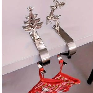 May include: Two silver Christmas stocking holders, one shaped like a tree and the other a reindeer. The holders are attached to a white surface, with red and white patterned stockings hanging below.