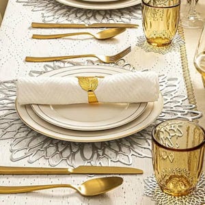 Könnte beinhalten: Elegante Tischdekoration mit goldfarbenem Besteck, weißen Tellern mit Goldrand und einer gerollten weißen Serviette, die von einem goldenen, blattförmigen Serviettenring gehalten wird. Bernsteinfarbene Gläser und dekorative Tischsets ergänzen das Arrangement.