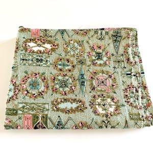 May include: A folded piece of fabric featuring a light green background with a repeating pattern of ornate, rectangular and circular frames. The designs include pink floral accents, dark green geometric lines, and small black details.