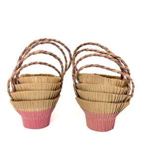 May include: Two stacks of cupcake liners, each featuring a pink base and a tan pleated upper section. Each stack is topped with a braided handle made of twisted pink and tan paper strips, creating a basket-like appearance for these baking cups.