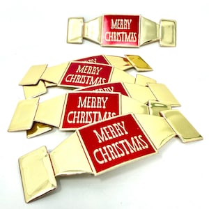 May include: A collection of five gold-toned metal decorative pieces shaped like wrapped candies. Each piece features a central red rectangular panel with the white text MERRY CHRISTMAS printed on it. The metallic finish is reflective and smooth.