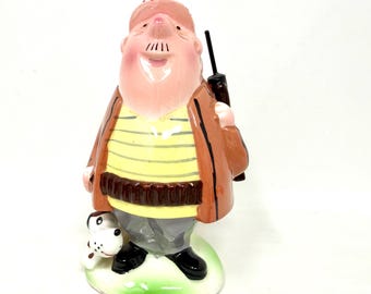 May include: A ceramic figurine of a hunter wearing a tan cap, brown jacket, yellow and white striped shirt, and gray trousers. The figure carries a black rifle over one shoulder and stands next to a small white and black spotted dog on a green base.