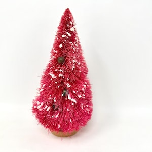 May include: A small, red bottle brush Christmas tree with white detailing. The tree has a wooden base and two small, dark-coloured ornaments. A festive decoration for the holiday season.
