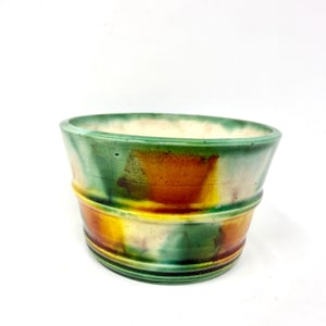 Puede incluir: Una maceta de cerámica con una parte superior ancha y abierta y una base redondeada. El exterior presenta una mezcla de tonos verdes, amarillos y naranjas, con un acabado texturizado y esmaltado. La maceta tiene un diseño simple pero elegante.