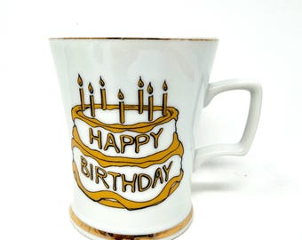 May include: White ceramic mug with a gold rim and handle. The mug features a birthday cake design with the words "HAPPY BIRTHDAY" in gold lettering. The cake has seven candles on top.