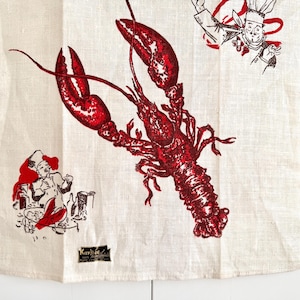 May include: Off-white linen kitchen towel with a large red lobster illustration. The design includes cartoon chefs and lobster claws. A small black label is at the bottom. The towel is likely for kitchen use.