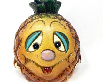 May include: A ceramic pineapple figurine with a smiling face. The pineapple has a yellow and brown body with a green top. The face features blue eyelids, black eyes, a purple nose, and a red tongue.