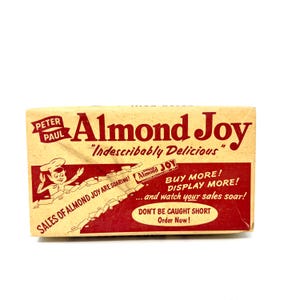 May include: A rectangular vintage Almond Joy candy display box with a cream and red colour scheme. The text reads Peter Paul Almond Joy, Indescribably Delicious, Sales of Almond Joy are soaring, Buy more, Display more, and watch your sales soar.