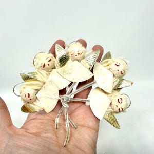 May include: A cluster of vintage Christmas angel ornaments. Each angel has a cream-colored face with red accents, gold wings, and a halo. The angels are attached to a silver pipe cleaner base.