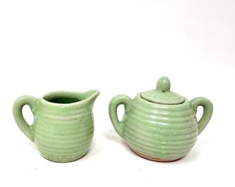 May include: A light green ceramic creamer and a matching lidded sugar bowl. Both pieces feature a horizontal ribbed texture, a glossy finish, and a rounded shape. The creamer has a single handle and a spout, while the sugar bowl has two handles.