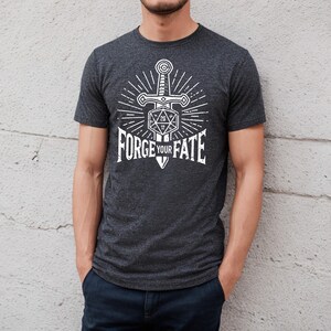 Peut inclure: Un t-shirt gris chiné foncé avec un graphique blanc d'une épée perçant un dé à vingt faces. Le graphique est entouré d'un motif de soleil. Le texte "FORGE YOUR FATE" est imprimé sous le graphique.