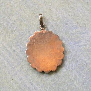 Puede incluir: Un colgante de metal de tono cobre, de forma redonda con un borde festoneado y una superficie lisa y plana. El colgante presenta un peque&ntilde;o lazo de metal circular en la parte superior, dise&ntilde;ado para sujetarse a una cadena o cord&oacute;n.