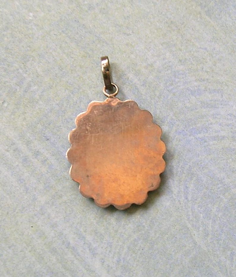 Puede incluir: Un colgante de metal de tono cobre, de forma redonda con un borde festoneado y una superficie lisa y plana. El colgante presenta un peque&ntilde;o lazo de metal circular en la parte superior, dise&ntilde;ado para sujetarse a una cadena o cord&oacute;n.