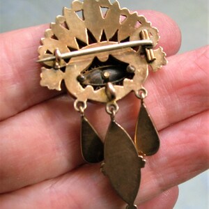 May include: A gold-coloured vintage brooch featuring a fan-shaped top and three hanging teardrop pendants. The brooch has an oval centre with a dark, possibly enamelled, design. The pin is held in a hand, showing the back of the brooch.