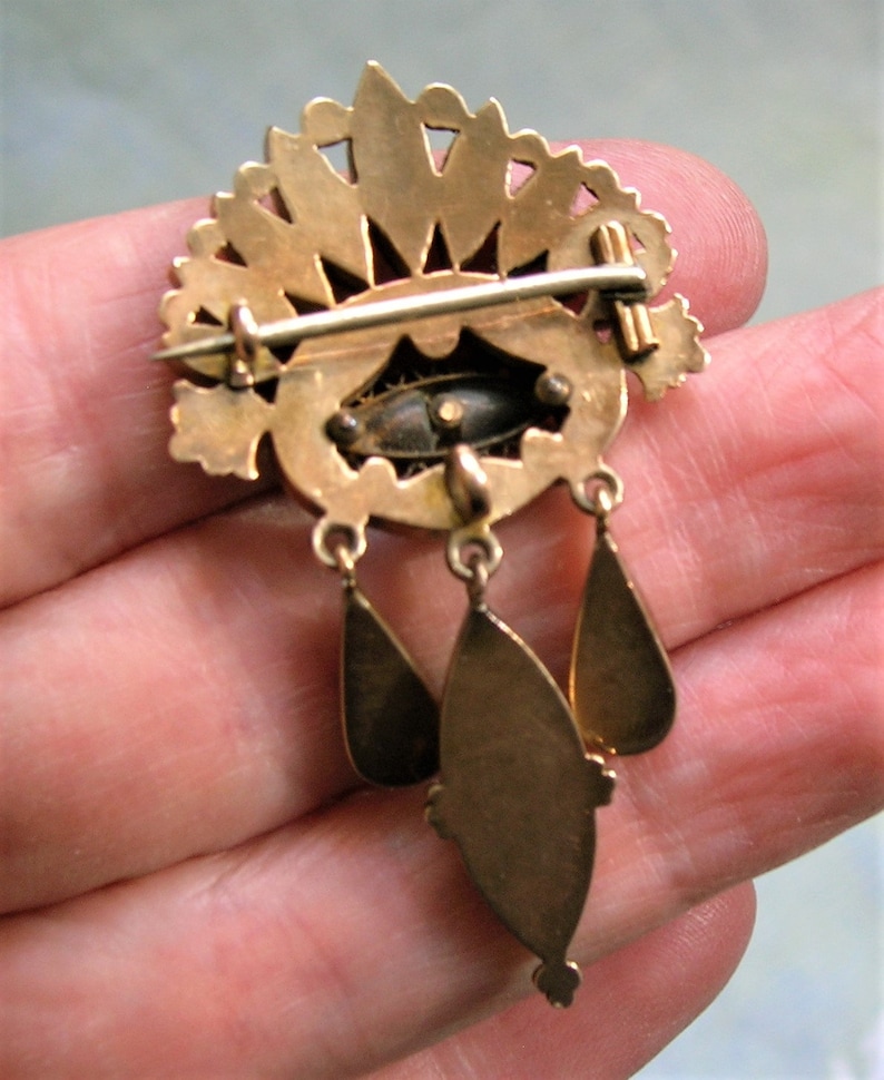May include: A gold-coloured vintage brooch featuring a fan-shaped top and three hanging teardrop pendants. The brooch has an oval centre with a dark, possibly enamelled, design. The pin is held in a hand, showing the back of the brooch.