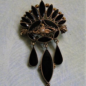 May include: An antique brooch with a gold-tone metal setting and black onyx stones. The brooch features a fan-shaped top with teardrop dangles. A small pearl is set in the centre of the brooch.