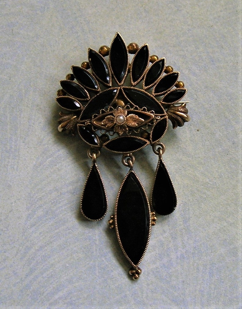 May include: An antique brooch with a gold-tone metal setting and black onyx stones. The brooch features a fan-shaped top with teardrop dangles. A small pearl is set in the centre of the brooch.
