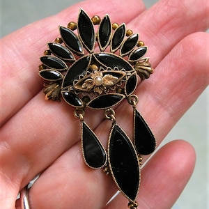 May include: An antique brooch with a gold-tone metal frame and black, teardrop-shaped accents. The brooch features a central oval design with a small pearl and floral details, and dangling teardrops.