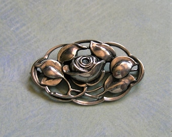 May include: A silver brooch with a rose and leaf design. The oval brooch features a detailed rose at its centre, surrounded by stylized leaves and an openwork frame. The metal has a polished finish.