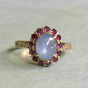 May include: A gold ring featuring a central oval cabochon light blue star sapphire surrounded by a halo of small round red rubies. The gold band is detailed with an engraved leaf pattern on both sides of the central gemstone setting.