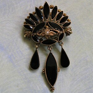 May include: A vintage gold-coloured brooch with black onyx detailing. The brooch has a fan-shaped top with teardrop and oval-shaped dangles. A small pearl is at the centre of the design. This brooch is an elegant piece of vintage jewellery.
