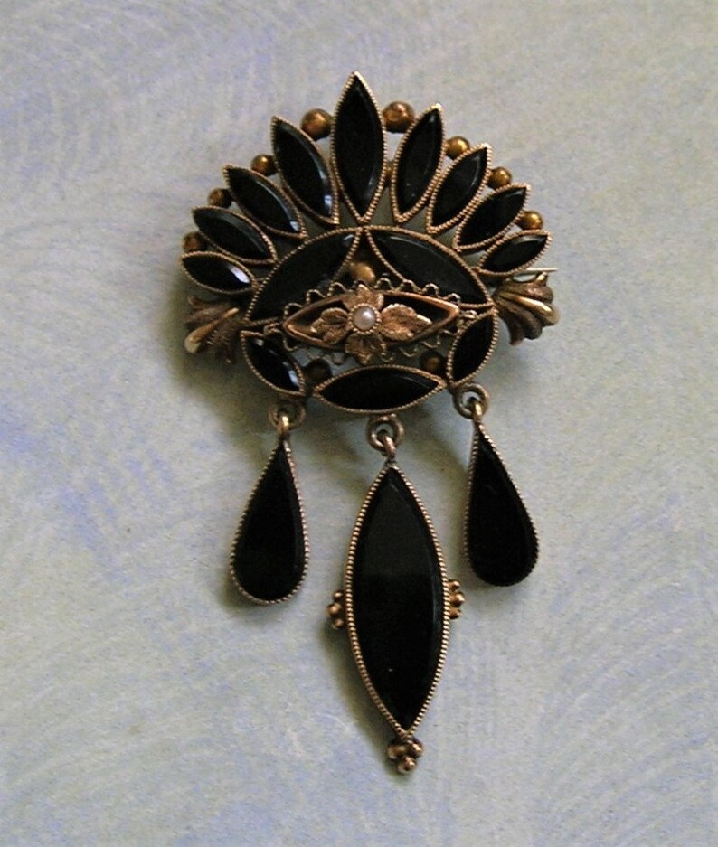 May include: A vintage gold-coloured brooch with black onyx detailing. The brooch has a fan-shaped top with teardrop and oval-shaped dangles. A small pearl is at the centre of the design. This brooch is an elegant piece of vintage jewellery.