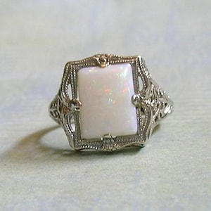 May include: A vintage-style ring featuring a rectangular, iridescent white opal stone set in an ornate, filigree silver-toned metal frame. The ring's design includes delicate scrollwork and decorative accents, showcasing a classic aesthetic.