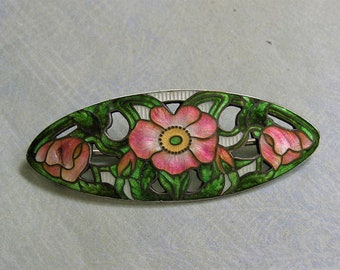 May include: Oval-shaped brooch with a floral design. The brooch features pink flowers with yellow centers and green leaves, set against a silver-colored metal. The design is detailed with enamel, creating a vibrant and artistic piece.