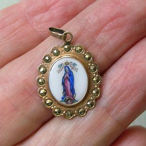 Puede incluir: Un colgante ovalado de tono dorado con una incrustaci&oacute;n central de esmalte blanco que muestra una figura con t&uacute;nicas azules y rosas siendo coronada por dos peque&ntilde;os &aacute;ngeles. El borde festoneado tiene piedras de marcasita gris oscuro.