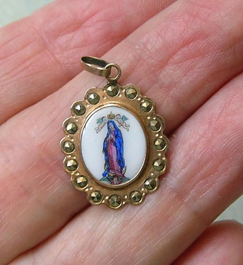 Puede incluir: Un colgante ovalado de tono dorado con una incrustaci&oacute;n central de esmalte blanco que muestra una figura con t&uacute;nicas azules y rosas siendo coronada por dos peque&ntilde;os &aacute;ngeles. El borde festoneado tiene piedras de marcasita gris oscuro.