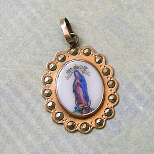 Puede incluir: Un colgante ovalado con una incrustaci&oacute;n central de esmalte blanco que representa una figura con t&uacute;nica azul y rosa, coronada por dos peque&ntilde;os &aacute;ngeles. El marco de metal dorado es festoneado y est&aacute; engastado con varias marcasitas facetadas.