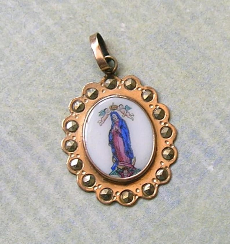 Puede incluir: Un colgante ovalado con una incrustaci&oacute;n central de esmalte blanco que representa una figura con t&uacute;nica azul y rosa, coronada por dos peque&ntilde;os &aacute;ngeles. El marco de metal dorado es festoneado y est&aacute; engastado con varias marcasitas facetadas.