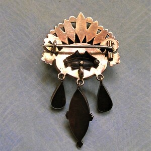 May include: A vintage gold-coloured brooch with a decorative fan-shaped top and three dangling teardrop pendants. The brooch has an oval centre with a pin closure on the back. The brooch is likely made of metal.