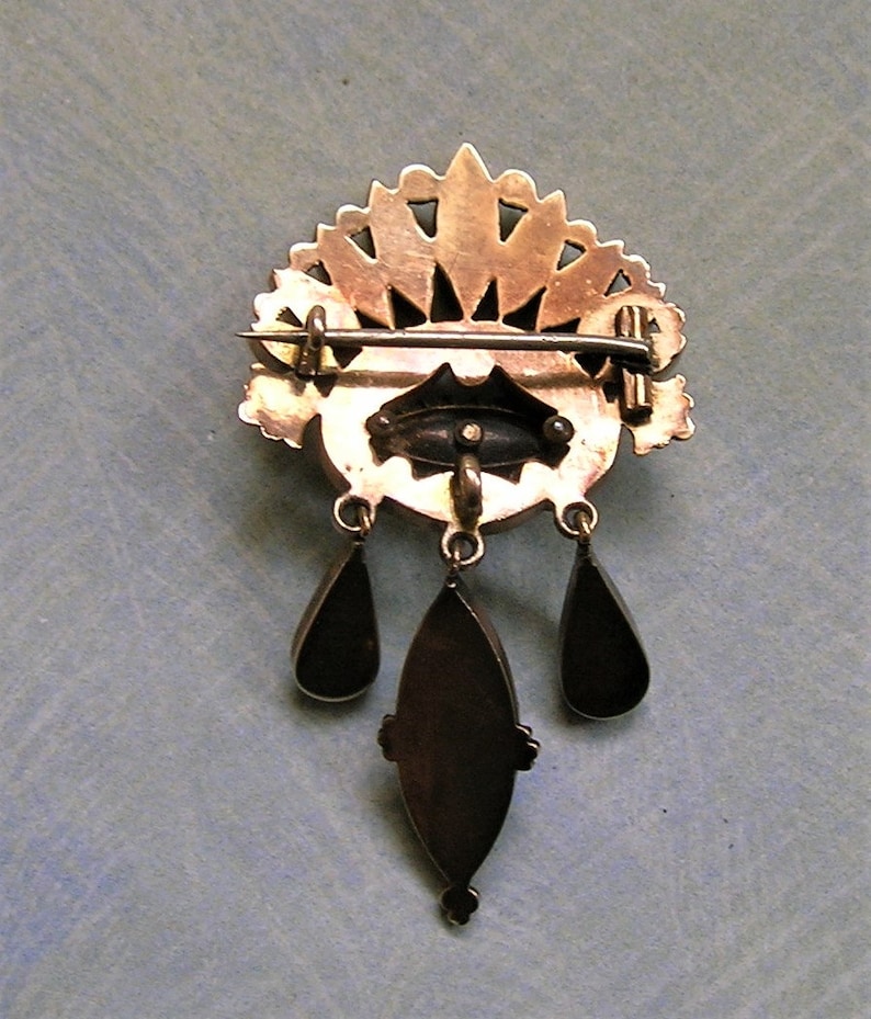 May include: A vintage gold-coloured brooch with a decorative fan-shaped top and three dangling teardrop pendants. The brooch has an oval centre with a pin closure on the back. The brooch is likely made of metal.