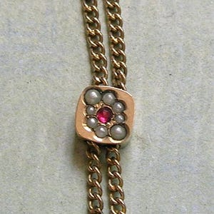 May include: A gold-toned chain necklace featuring a square-shaped slider pendant. The pendant is set with a central round red stone surrounded by eight small white pearls arranged in a cluster pattern, suspended on a double-strand gold chain.
