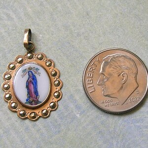 Puede incluir: Un colgante ovalado con una incrustaci&oacute;n central de esmalte blanco que muestra una figura con t&uacute;nicas azules y rosas, rodeado por un marco dorado festoneado con piedras de marcasita, junto a una moneda de diez centavos de EE. UU. de 2022.