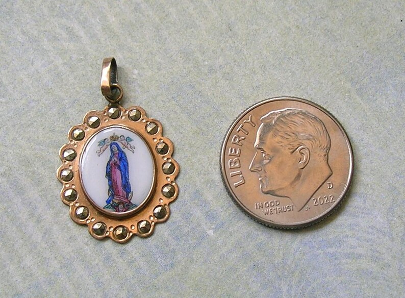 Puede incluir: Un colgante ovalado con una incrustaci&oacute;n central de esmalte blanco que muestra una figura con t&uacute;nicas azules y rosas, rodeado por un marco dorado festoneado con piedras de marcasita, junto a una moneda de diez centavos de EE. UU. de 2022.