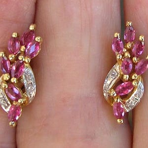 May include: A pair of gold earrings featuring marquise-cut pink rubies arranged in a cluster above a curved gold band set with small, clear, rectangular-cut diamonds. The design incorporates multiple pink stones and metallic gold accents.