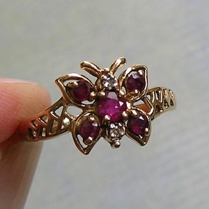 May include: A gold-toned ring featuring a butterfly-shaped centerpiece. The butterfly is adorned with four deep red circular gemstones on the wings, a central oval red gemstone, and two small clear white stones positioned at the top and bottom.