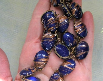 May include: A vintage necklace featuring dark blue oval-shaped beads with gold-colored swirl patterns. The beads are linked together with gold-toned metal connectors. The necklace is a striking piece of jewelry.