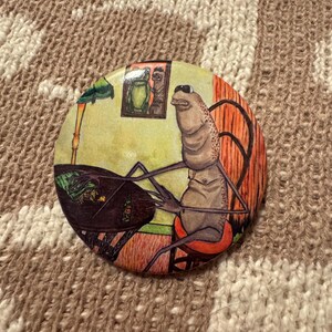 May include: Round button with a whimsical illustration of a creature seated at a table. The creature has a light brown body, large eyes, and is seated in a red chair. The background features a framed picture and a green lamp.