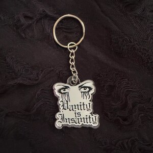 May include: A silver keychain with a black and white acrylic charm. The charm features a pair of eyes with tears and the text "Vanity is Insanity".