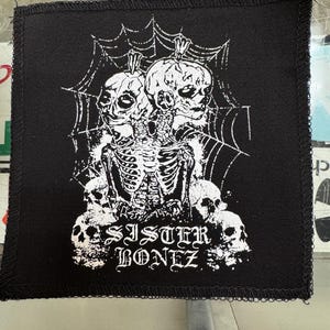 May include: Black square patch with a white graphic of two conjoined skulls, a rib cage, and spiderwebs. The text "SISTER BONEZ" is below the graphic. The patch has a black border.
