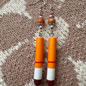 Op de afbeelding: Een paar handgemaakte oorbellen met een uniek ontwerp. De oorbellen hebben oranje cilinders met witte en kastanjebruine accenten, bekroond met decoratieve kralen in oranje, bruin en wit. Ze hangen aan zilverkleurige haken.