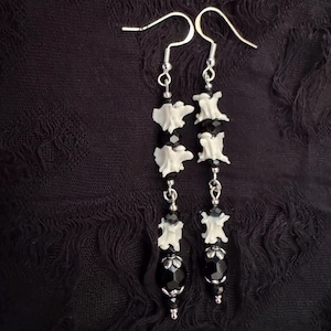 May include: A pair of dangle earrings with a gothic aesthetic. Each earring features white bone-shaped beads, black faceted beads, and silver-colored metal accents. The earrings hang from silver-colored ear wires against a dark background.